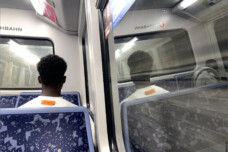 Fotografie von Hilmar Sundermann. Rückansicht eines jungen schwarzen Mannes mit kurzer Afrofrisur und weißem T-Shirt mit orangefarbenem Etikett auf dem Rücken. Er sitzt in einer U-Bahn und spiegelt sich im Fenster wie auch eine weiter entfernt sitzende Person.