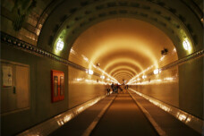 Fotografie von Hilmar Sundermann im St. Pauli Elbtunnel. Blick in eine der Röhren mit gewölbter Decke und Kacheln an den Wänden mit sternförmig aus dem Fluchtpunkt laufenden Lichtspuren. Eine Menschengruppe weiter hinten im Tunnel. Ein roter Notfallschrank an der Wand im Vordergrund.