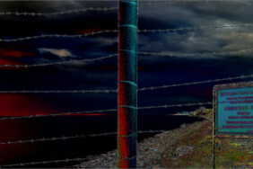 Fotografie von Hilmar Sundermann in surrealer Bearbeitung mit kräftigen Farben. Blick durch einen Stacheldrahtzaun über den Strand zum schwarz-grauen Himmel mit dunklen Rottönen; seitlich direkt hinter dem Zaun ein Gefahren-Hinweisschild, dahinter das Kliff.