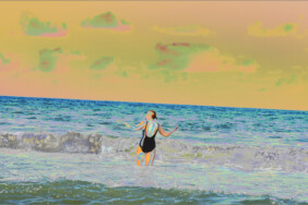 Fotografie von Hilmar Sundermann in surrealer Bearbeitung mit matt-leuchtenden Gelb- Blau- und Türkistönen. Eine Badende im schwarzen Badeanzug steht mit seitlich leicht erhobenen Armen im flachen Wasser am Ostseestrand und blickt zum schrägen Horizont.