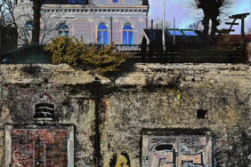 Fotografie von Hilmar Sundermann in surrealer Bearbeitung mit matten Grau-, Rot- und Blautönen. Verwitterte Mauer am Hamburger Elbstrand bei Övelgönne mit Graffiti, unter anderem ein Frauenkopf, und eingelassenen, mit vielen Wörtern beschriebenen Türen; darüber ein älteres Gebäude mit Ecktürmchen.