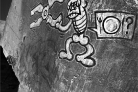 Fotografie von Hilmar Sundermann in Schwarz-Weiß-Bearbeitung. Vor schwarzem Hintergrund ein grauer Bunker auf dem Dünensand mit weißem Graffito einer stilisierten, skelettartigen Figur mit übergroßer Zange in den Händen, daneben eine rechteckige Sprechblase mit Teller, Besteck und einem Fragezeichen; unten auf dem Bunker der Schatten des Fotografen mit zum Kopf hin angewinkeltem Arm und darunter seitlich hängender Tasche.