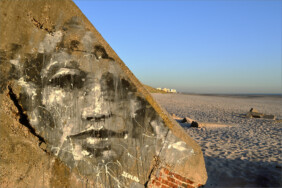 Fotografie von Hilmar Sundermann. Im Vordergrund ein massiver bräunlicher Betonbunker am Strand, darauf ein großflächiges, schwarz-weißes Porträt-Graffito mit Blick über den weiten Sandstrand; am Horizont die weißen Hochhäuser von Hardelot-Plage, darüber ein klarer blauer Himmel.