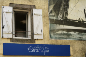 Fotografie von Hilmar Sundermann in überwiegend matten Farben. Gezeigt wird ein Ausschnitt der verwitterten gelblichen Fassade eines Geschäftshauses im bretonischen Douarnenez; daran das großformatige Foto eines Segelboots in Nahaufnahme mit mehreren Personen an Bord; daneben gibt ein geöffnetes Fenster mit weißen Fensterläden den Blick in ein dunkles Zimmer frei, in dem eine futuristisch wirkende Lampe mit rundgebogenen Neonröhren hängt; unter Fenster und Foto ein blaues Schild mit der weißen Aufschrift „atelier d'art Ceramique“.